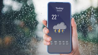 雨に覆われた窓の前で、雷雨が予想されている天気予報アプリを表示している携帯電話