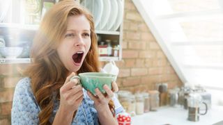 赤い髪の女性がキッチンに立ち、緑茶のカップを持ちながらあくびをしている